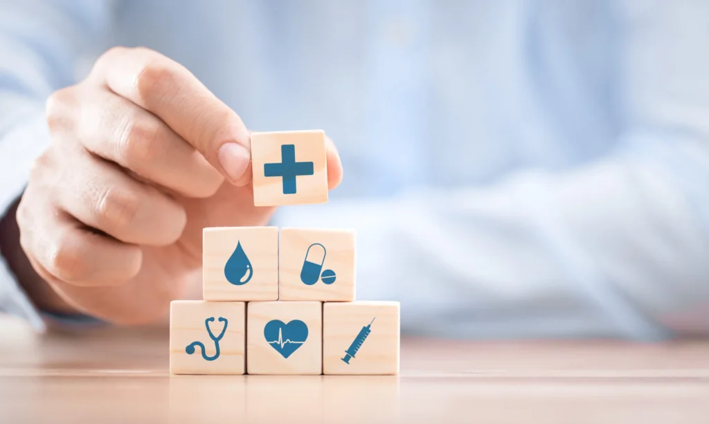 Hand stacking wooden blocks with medical icons, including a cross, stethoscope, pills, syringe, and heart, representing medication management.